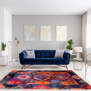 Op de afbeelding: Een blauwe fluwelen bank met twee kussens staat in een woonkamer met een kleurrijk tapijt met patronen, een gouden staande lamp en een gouden metalen stoel. De muren zijn grijs geverfd en er hangen drie abstracte schilderijen aan de muur.