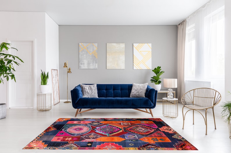 Op de afbeelding: Een blauwe fluwelen bank met twee kussens staat in een woonkamer met een kleurrijk tapijt met patronen, een gouden staande lamp en een gouden metalen stoel. De muren zijn grijs geverfd en er hangen drie abstracte schilderijen aan de muur.