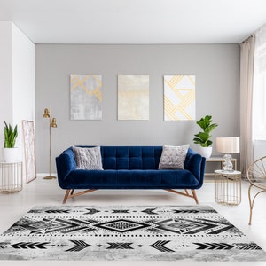 K&ouml;nnte beinhalten: Ein blaues Samtsofa mit zwei Kissen steht in einem Wohnzimmer mit einem geometrischen Teppich in Schwarzwei&szlig;. Der Raum hat wei&szlig;e W&auml;nde, goldene Akzente und ein gro&szlig;es Fenster mit durchsichtigen Vorh&auml;ngen.