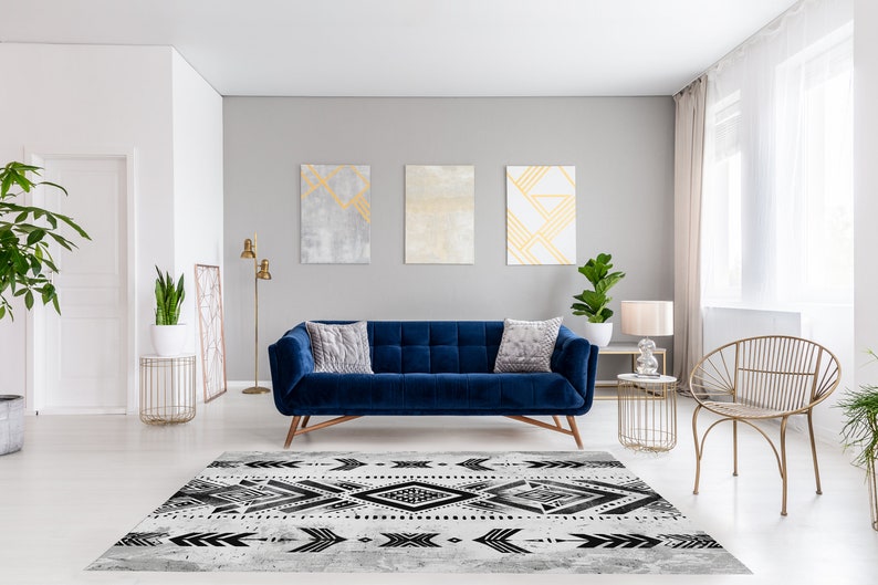 K&ouml;nnte beinhalten: Ein blaues Samtsofa mit zwei Kissen steht in einem Wohnzimmer mit einem geometrischen Teppich in Schwarzwei&szlig;. Der Raum hat wei&szlig;e W&auml;nde, goldene Akzente und ein gro&szlig;es Fenster mit durchsichtigen Vorh&auml;ngen.