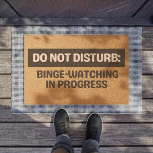 May include: A brown doormat with the text "DO NOT DISTURB: BINGE-WATCHING IN PROGRESS" on it. The doormat is on a wooden floor.