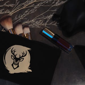May include: Black makeup bag with a gold deer logo, containing makeup brushes. A tube of dark red lipstick sits next to the bag. The items are arranged on a dark surface with decorative sprigs.