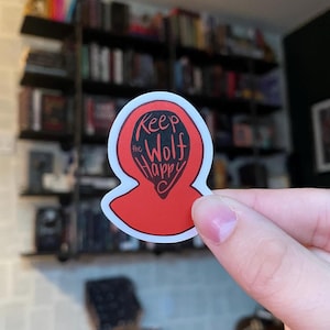 Puede incluir: Pegatina roja y blanca con contorno negro. La pegatina tiene forma de figura encapuchada y dice "Keep the Wolf Happy" en texto negro.