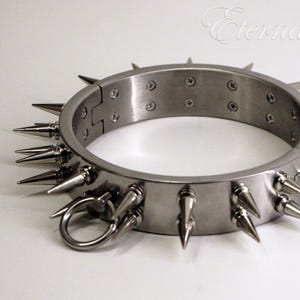 Può includere: Un collare in metallo color argento con affilate punte coniche. Il collare ha un design a cerniera con un anello ed è fissato con viti. Il testo "Eternal Fetish" è visibile nell'angolo in alto a destra.