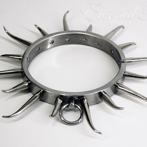 Könnte beinhalten: Ein silberfarbener Metallkragen mit nach außen gerichteten Stacheln. Der Kragen hat ein kreisförmiges Design mit einem Ring an der Unterseite. Der Text "Eternal Fetish" ist oben rechts zu sehen.
