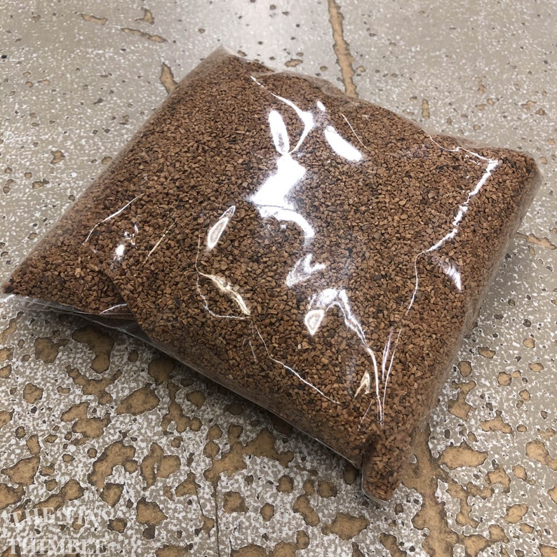 Pu&ograve; includere: Due sacchetti di plastica trasparente pieni di piccoli pezzi di sughero marrone di forma irregolare. I sacchetti sono leggermente trasparenti, permettendo di vedere il materiale di sughero all'interno. I sacchetti sono appoggiati su una superficie strutturata di colore chiaro.