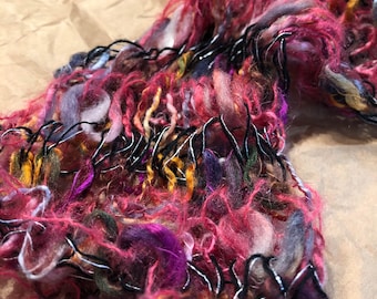 May include: A close-up of a pink, purple, and gray yarn scarf with black wire accents. The scarf is made of a soft, fluffy yarn and has a unique, textured look.
