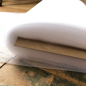 Puede incluir: Tela de tul blanco enrollada en un tubo de cartón marrón. El tul es suave y delicado, perfecto para manualidades o decoración.