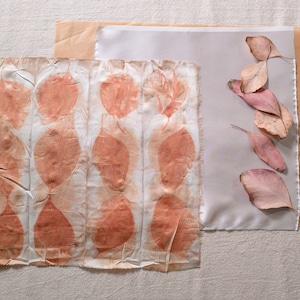 May include: A white fabric with a pattern of orange leaf prints. The fabric is laid out on a beige surface next to a piece of brown paper, a wooden tool, and a rubber band. There are also several dried leaves on the brown paper.
