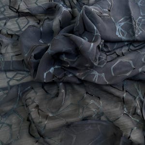 May include: Dark gray fabric with a geometric pattern. The fabric is draped and folded, creating shadows and texture. The pattern appears to be a series of interconnected shapes in a lighter shade of gray. The fabric is semi-transparent. The text "THE TIN THIMBLE" is visible.