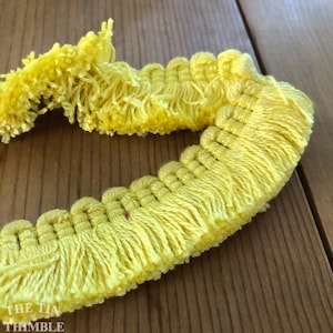 May include: Close-up of a length of bright yellow fringe trim. The trim has a looped edge with long, soft tassels. The text "THE TIN THIMBLE" is visible in the lower left corner. The trim is lying on a wooden surface.