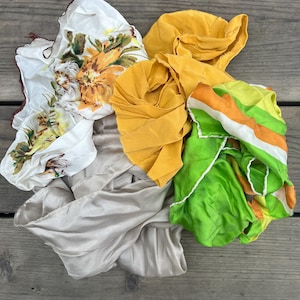 May include: A collection of four silk scarves in various colors and patterns. The scarves are folded and piled on top of each other. The scarves are a light beige, a bright yellow, a white with brown and yellow floral print, and a green with orange and white stripes.