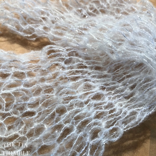May include: Close-up of a white, open-weave yarn with silver metallic threads. The yarn has a soft, fluffy texture and is slightly translucent. The image is well-lit, highlighting the delicate structure of the fiber. The Tin Thimble logo is in the bottom left corner.