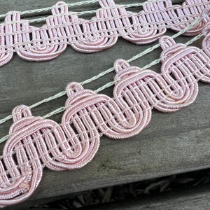 May include: Pink scalloped trim with a woven texture. The trim is made of a soft, pliable material and has a delicate, feminine look.