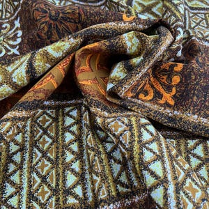 May include: Close-up of a patterned fabric with a vintage aesthetic. The design features geometric shapes, floral motifs, and a mix of colors including brown, orange, and cream. The fabric is draped, showing its texture and folds.