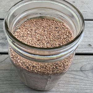 Pu&ograve; includere: Un barattolo di vetro trasparente pieno di piccoli pezzi di materiale marrone di forma irregolare. Il barattolo ha un'ampia apertura e un anello di metallo sulla parte superiore. Il testo "THE TIN THIMBLE" &egrave; visibile in basso a sinistra.