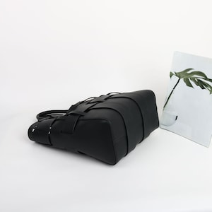 Puede incluir: Un bolso tote de cuero negro con un dise&ntilde;o tejido. El bolso tiene forma rectangular con un asa y un patr&oacute;n tejido &uacute;nico en la parte delantera. El bolso est&aacute; sobre un fondo blanco.