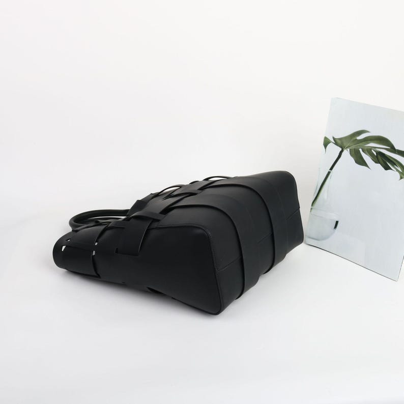 Puede incluir: Un bolso tote de cuero negro con un dise&ntilde;o tejido. El bolso tiene forma rectangular con un asa y un patr&oacute;n tejido &uacute;nico en la parte delantera. El bolso est&aacute; sobre un fondo blanco.