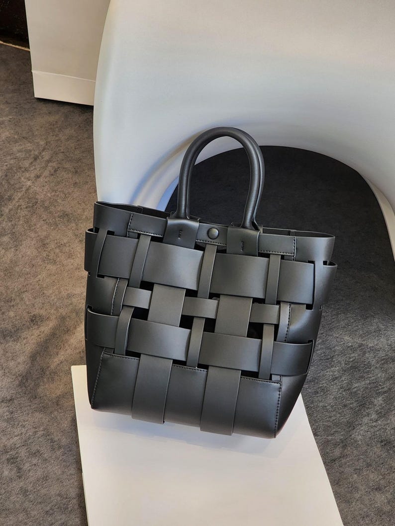 Puede incluir: Un bolso tote negro con un dise&ntilde;o tejido. El bolso tiene una forma estructurada con un asa superior y un cierre de bot&oacute;n. El patr&oacute;n tejido crea una textura &uacute;nica. El bolso est&aacute; sobre una superficie blanca.