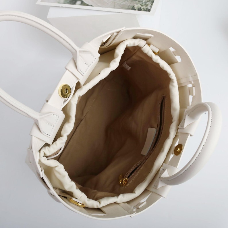 Puede incluir: Bolso tote color crema con dise&ntilde;o tejido y dos asas. El interior cuenta con un cierre de cord&oacute;n y un bolsillo con cremallera. El bolso tiene herrajes dorados y un forro de tono neutro.