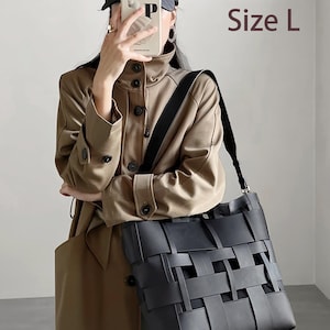 Puede incluir: Un bolso tote negro tejido con una correa para el hombro. El bolso lo sostiene una persona que lleva una gabardina color beige y una gorra negra. El texto "Size L" es visible.