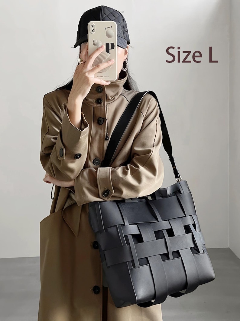 Puede incluir: Un bolso tote negro tejido con una correa para el hombro. El bolso lo sostiene una persona que lleva una gabardina color beige y una gorra negra. El texto "Size L" es visible.