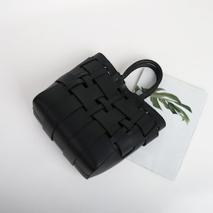 Puede incluir: Un bolso tote negro tejido con forma rectangular y un asa. La bolsa est&aacute; hecha de tiras de material entrelazadas, creando un dise&ntilde;o similar a una cesta. El asa tiene forma redondeada.