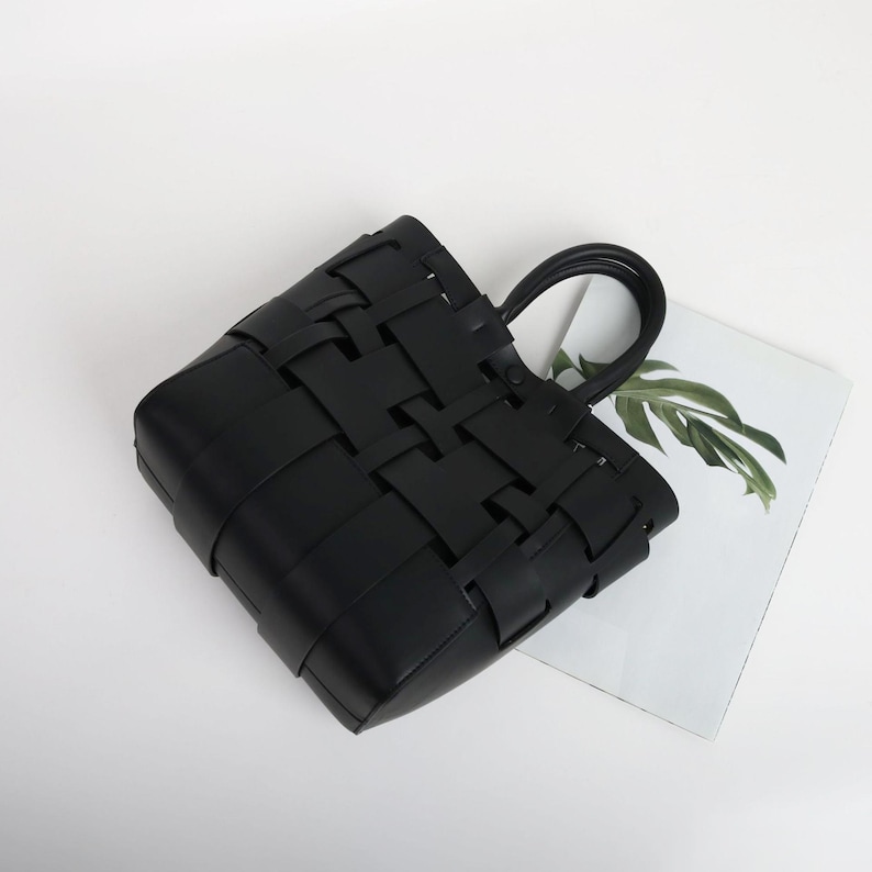 Puede incluir: Un bolso tote negro tejido con forma rectangular y un asa. La bolsa est&aacute; hecha de tiras de material entrelazadas, creando un dise&ntilde;o similar a una cesta. El asa tiene forma redondeada.