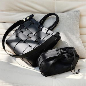 Puede incluir: Un bolso tote de cuero negro tejido con un asa superior y una correa para el hombro desmontable. Una peque&ntilde;a bolsa negra con cord&oacute;n se encuentra junto al bolso tote. El bolso tiene una hebilla dorada.