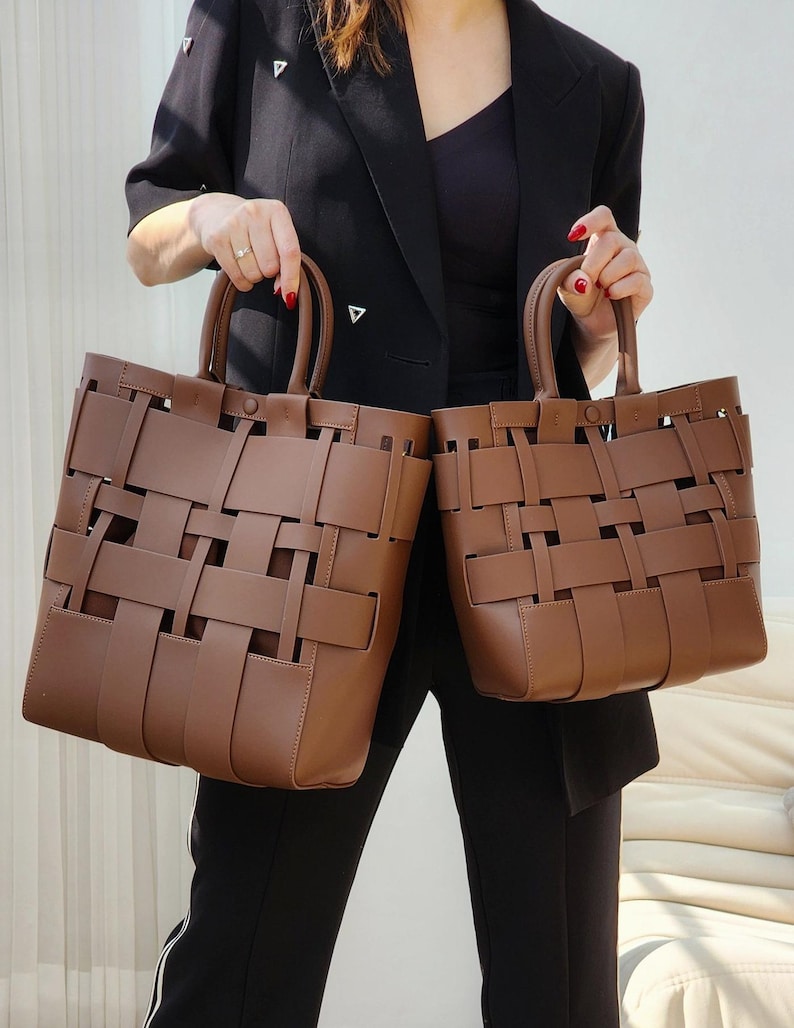 Puede incluir: Dos bolsos tote marrones tejidos con asas superiores. Las bolsas tienen forma rectangular y un dise&ntilde;o de tejido de cesta. Las bolsas son sostenidas por una persona que lleva una blazer negra y pantalones negros.