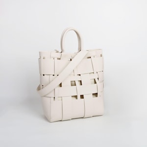 Puede incluir: Bolso tote tejido de color crema con un asa superior y una correa para el hombro desmontable. El bolso presenta un dise&ntilde;o de tejido de cesta, con tiras rectangulares que crean un patr&oacute;n abierto. El bolso es de color neutro y parece ser de cuero.