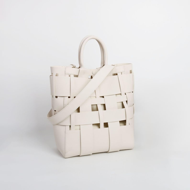 Puede incluir: Bolso tote tejido de color crema con un asa superior y una correa para el hombro desmontable. El bolso presenta un dise&ntilde;o de tejido de cesta, con tiras rectangulares que crean un patr&oacute;n abierto. El bolso es de color neutro y parece ser de cuero.