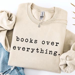 May include: A sand-colored sweatshirt with the text "books over everything" in black. The sweatshirt is folded with a pair of blue jeans and a necklace. The word "SAND" is printed on a white label.