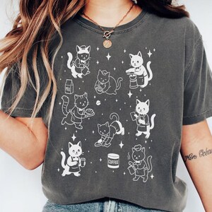 May include: Dark grey t-shirt featuring a repeating pattern of white cartoon cats in various poses, some holding food and drinks. The cats are depicted with chef hats and aprons. The shirt is a comfortable, casual style.
