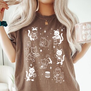May include: A brown t-shirt featuring a repeating pattern of white cartoon cats in various poses, some holding coffee cups and milk cartons. The shirt is worn by a person holding a pink mug with the text "Cool Moms Club".