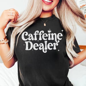 May include: Black t-shirt with the text "Caffeine Dealer" in white, stylized font. The design includes a small coffee cup icon above the word "Dealer" and star accents. The shirt is a solid black color.