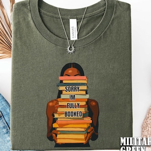 以下が含まれることがあります： ミリタリーグリーンのTシャツ。本を抱える人物のグラフィックが描かれています。本には「SORRY IM FULLY BOOKED」と書かれています。太陽のペンダントが付いたシルバーのネックレスが見えます。Tシャツは折りたたまれています。