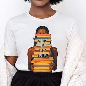 以下が含まれることがあります： 本を抱える人のグラフィックがプリントされた白いTシャツ。「SORRY IM FULLY BOOKED」の文字が本にプリントされています。その人は肌が黒く、黒髪です。このシャツは黒いショートパンツと白いカーディガンと組み合わせています。