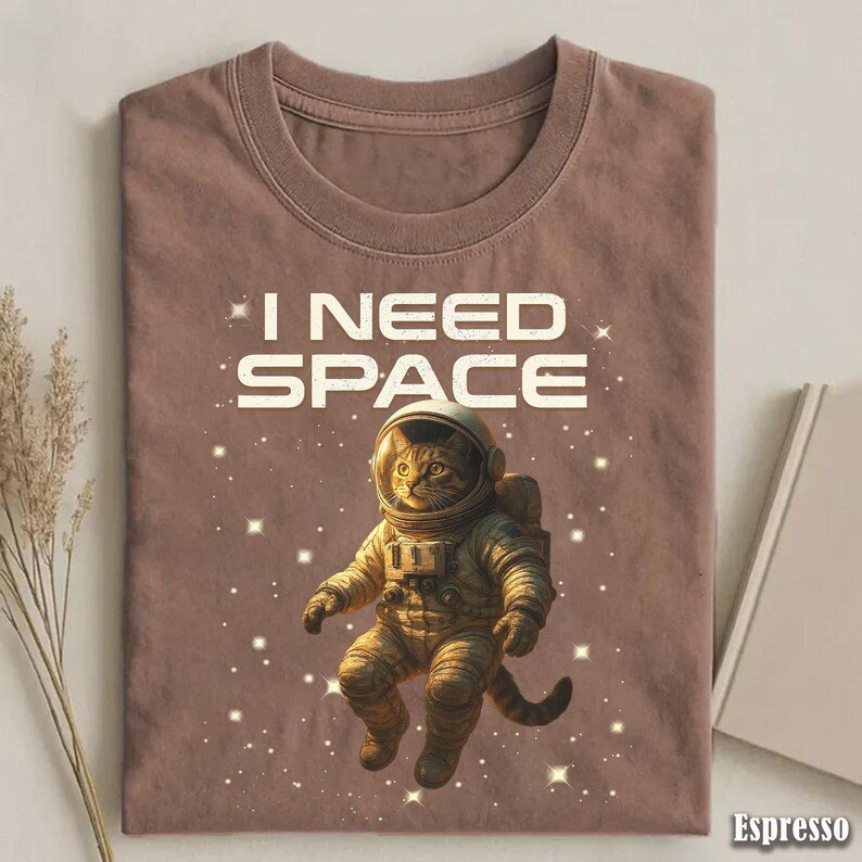 May include: Espresso-colored t-shirt featuring a cat astronaut floating in space, surrounded by stars. The text "I NEED SPACE" is printed above the cat. The shirt is made of a soft material.