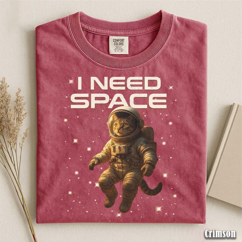 May include: Crimson-colored t-shirt with the text "I NEED SPACE" above an illustration of a cat in an astronaut suit, surrounded by stars. The shirt is made of Comfort Colors material.