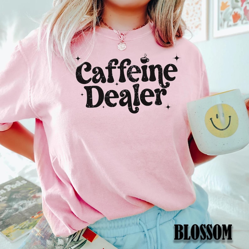 May include: A light pink sweatshirt with the text "Caffeine Dealer" in black, along with a coffee cup graphic. A white mug with a yellow smiley face is held in the hand. The word "BLOSSOM" is printed at the bottom.