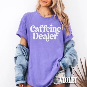 May include: A violet-colored t-shirt with the text "Caffeine Dealer" in white, featuring a coffee cup graphic. The word "VIOLET" is printed at the bottom right corner. The shirt has rolled-up sleeves.