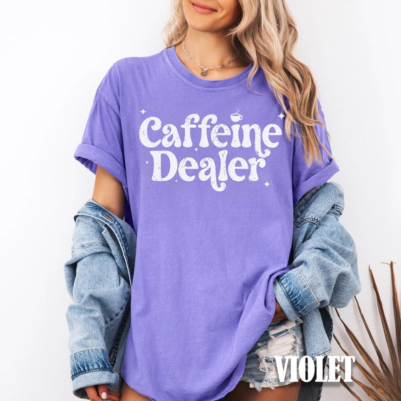 May include: A violet-colored t-shirt with the text "Caffeine Dealer" in white, featuring a coffee cup graphic. The word "VIOLET" is printed at the bottom right corner. The shirt has rolled-up sleeves.