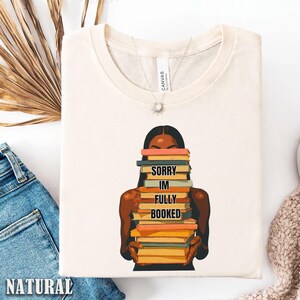 以下が含まれることがあります： クリーム色のTシャツ。本を抱える人物のグラフィックが特徴です。本は様々な色で、テキスト「SORRY IM FULLY BOOKED」が本に印刷されています。柔らかい素材で作られたシャツです。