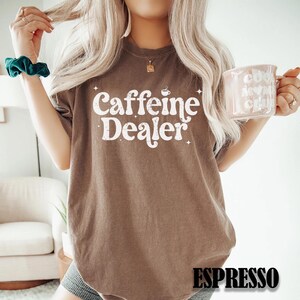 May include: Brown t-shirt with the text "Caffeine Dealer" in white, with a coffee cup graphic. The word "Espresso" is printed at the bottom. The person is holding a pink mug.