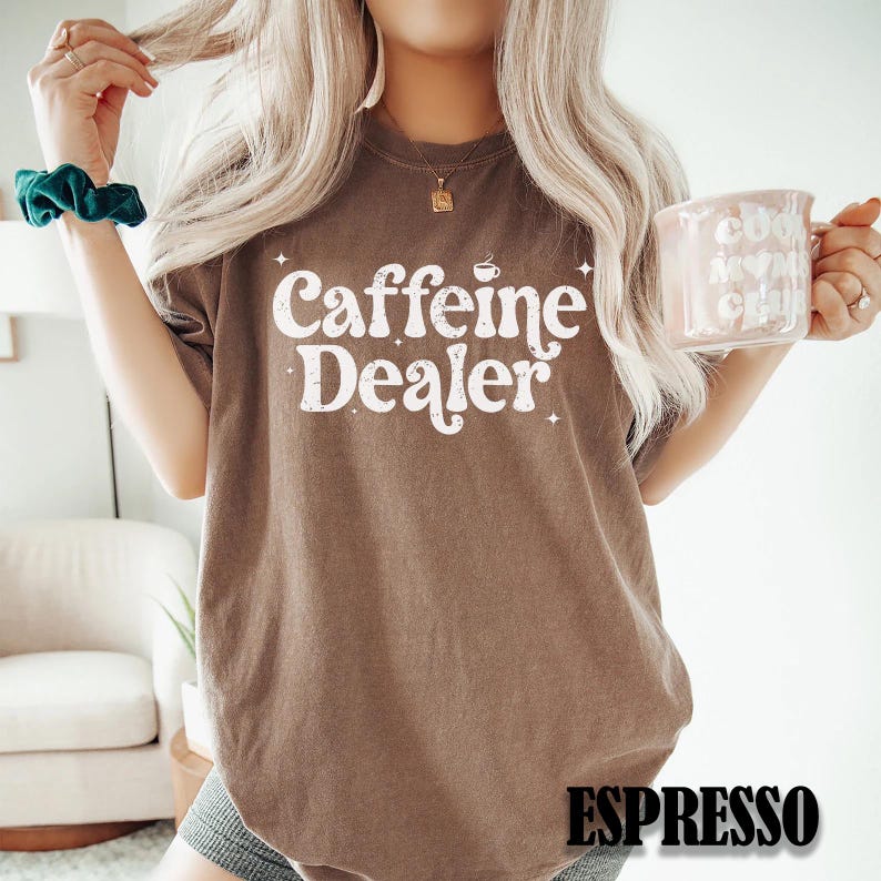 May include: Brown t-shirt with the text "Caffeine Dealer" in white, with a coffee cup graphic. The word "Espresso" is printed at the bottom. The person is holding a pink mug.