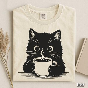 Peut inclure: T-shirt ivoire avec un chat noir tenant une tasse de caf&eacute;. Le chat a de grands yeux et des moustaches, avec de la vapeur qui s'&eacute;chappe de la tasse. Le t-shirt a un col rond et une &eacute;tiquette Comfort Colors.