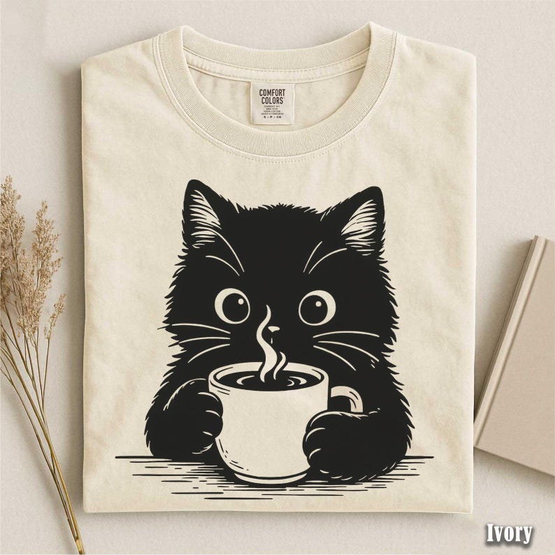 Peut inclure: T-shirt ivoire avec un chat noir tenant une tasse de caf&eacute;. Le chat a de grands yeux et des moustaches, avec de la vapeur qui s'&eacute;chappe de la tasse. Le t-shirt a un col rond et une &eacute;tiquette Comfort Colors.