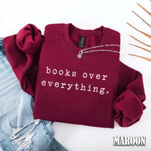 May include: A maroon sweatshirt with the text "books over everything" in white. A silver necklace with a circular pendant rests on the shirt. The sweatshirt is folded over a pair of blue jeans.