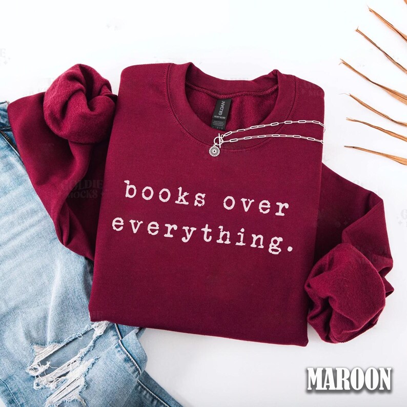 May include: A maroon sweatshirt with the text "books over everything" in white. A silver necklace with a circular pendant rests on the shirt. The sweatshirt is folded over a pair of blue jeans.